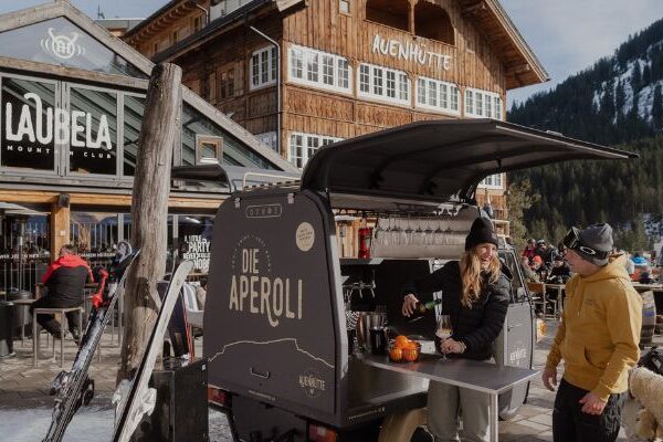 Aperol-Bar vor der Auenhütte an der Ifenbahn im Kleinwalsertal mit Skifahrern auf der Sonnenterrasse