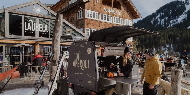 Aperol-Bar vor der Auenhütte an der Ifenbahn im Kleinwalsertal mit Skifahrern auf der Sonnenterrasse